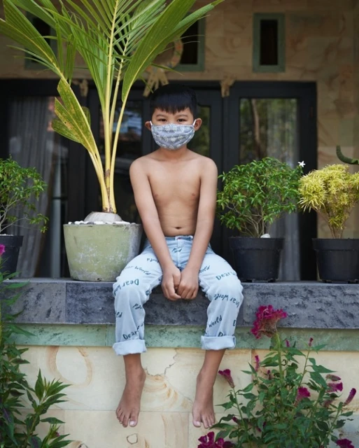 Young child with a face mask sits on a ledge surrounded by potted plants outside a house, bathed in soft daylight, in a medium portrait shot.