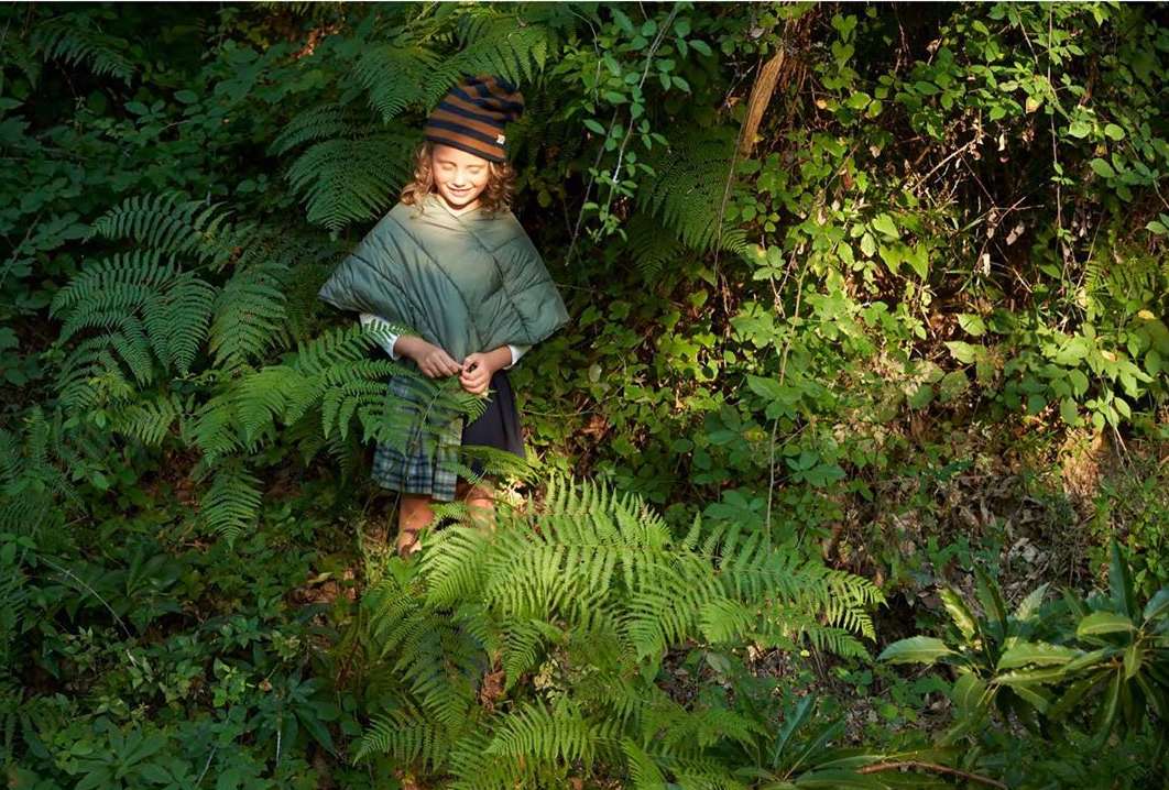 A girl in a striped hat and poncho stands among lush green foliage, ferns, and sunlight in a natural outdoor environment.