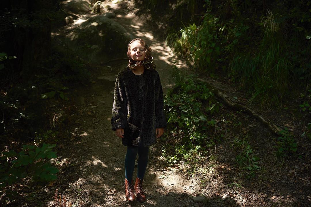 A young girl stands on a path in a sun-dappled forest wearing a faux-fur coat, leggings, and boots in a full shot.