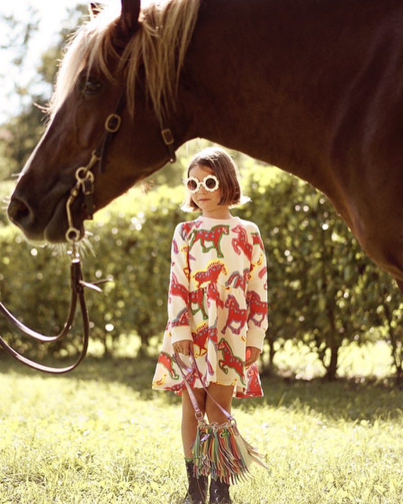 Child in horse-print dress stands with horse in green field wearing flower sunglasses and carrying a colorful fringe purse.