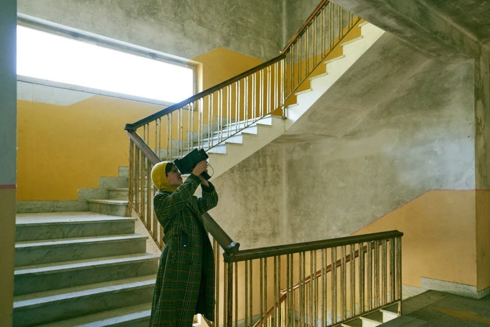 Eye-level shot in an indoor environment of a woman in a coat and headscarf holding a camera, framed by a staircase with yellow walls.