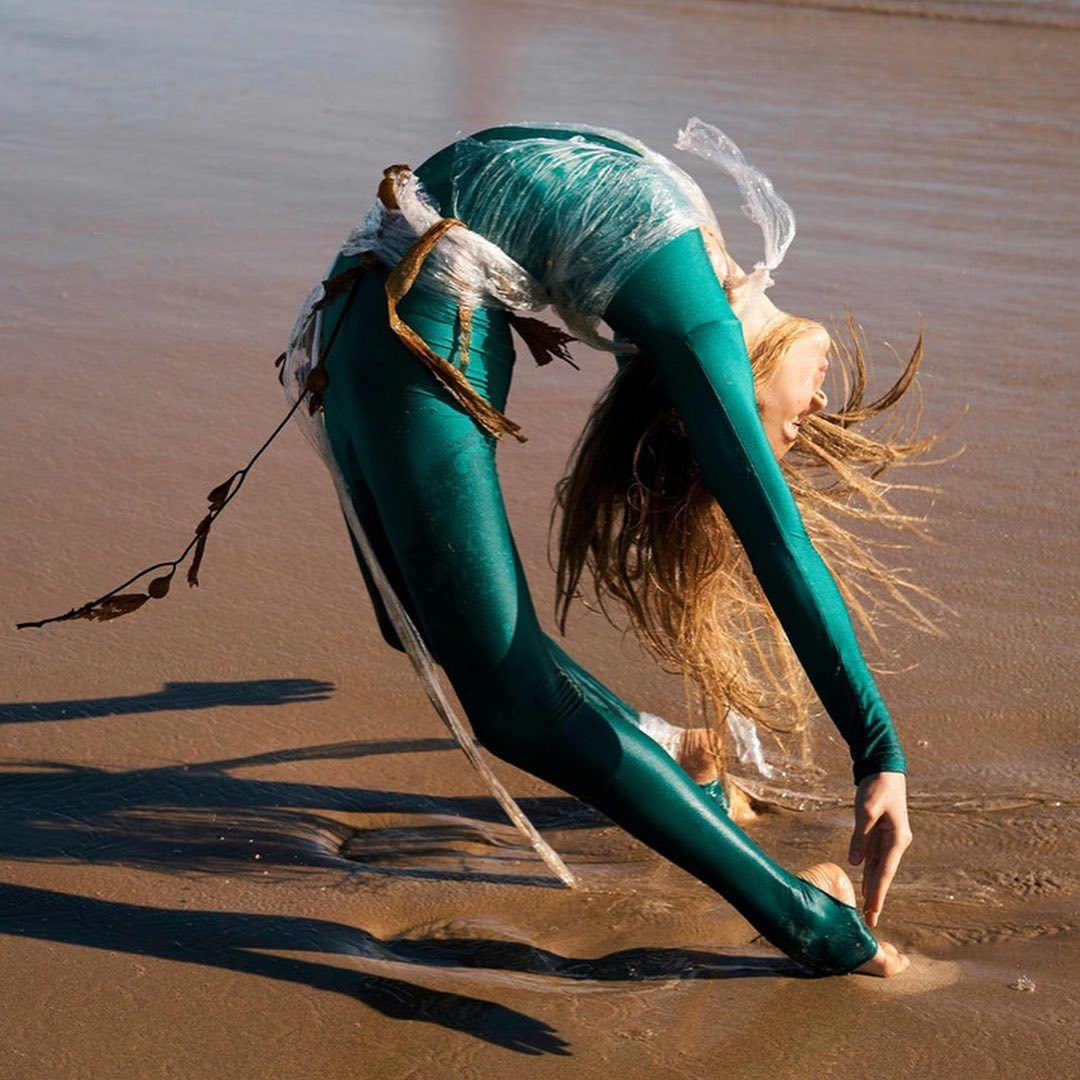 A woman in a green bodysuit stands on a sandy beach, bending backward with her hands touching the sand. She is partially wrapped in plastic.