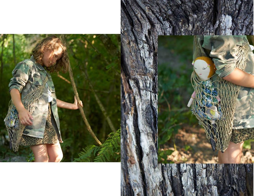 Young girl in camouflage jacket and leopard print skirt carrying a doll in a net bag, standing outdoors with a tree trunk.