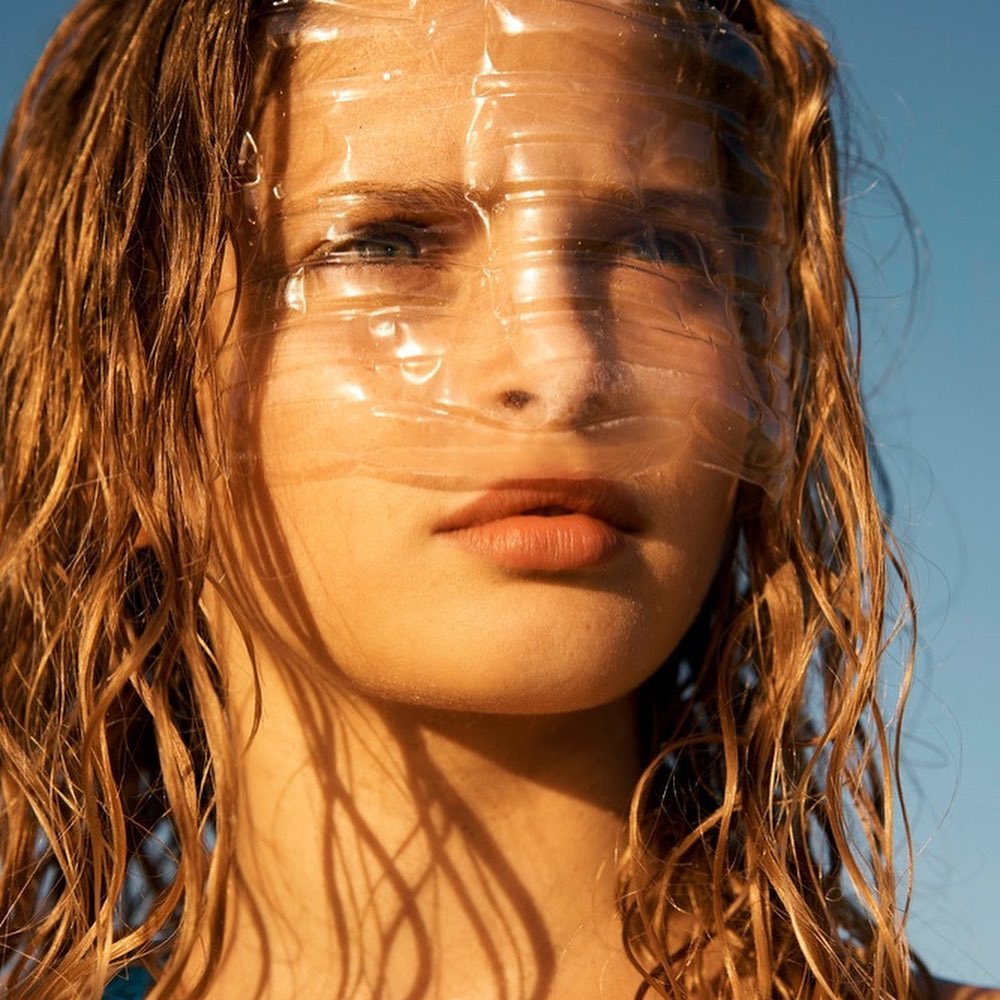 Portrait of a blonde adult female with wet hair and a transparent plastic wrap on her face against a clear blue sky in natural light.