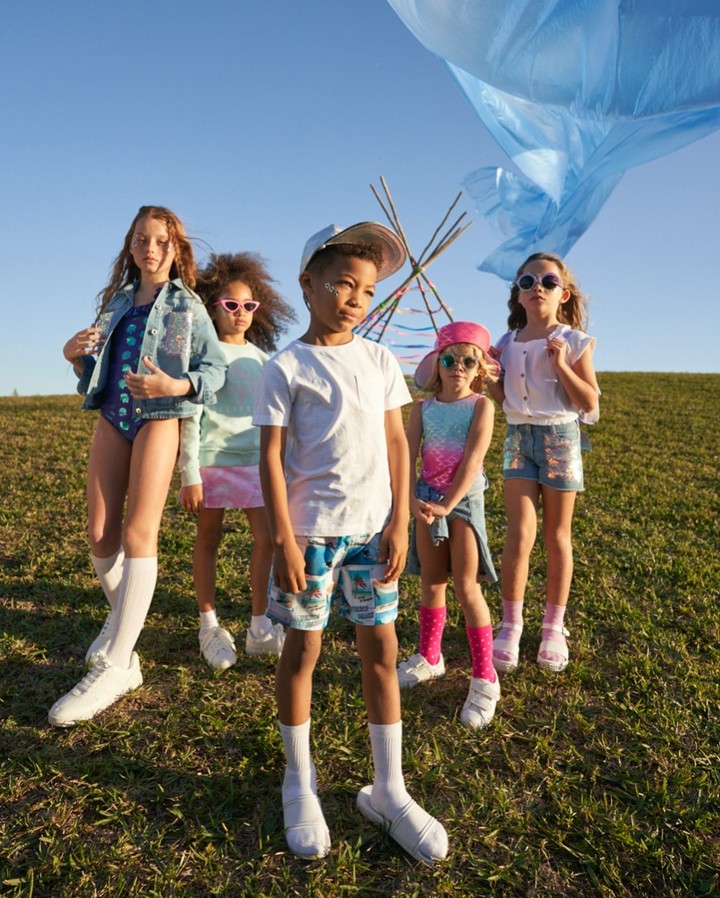 Group of five kids in stylish summer clothing posing on grassy hill with blue sky. One boy in foreground wearing a white t-shirt and patterned shorts.