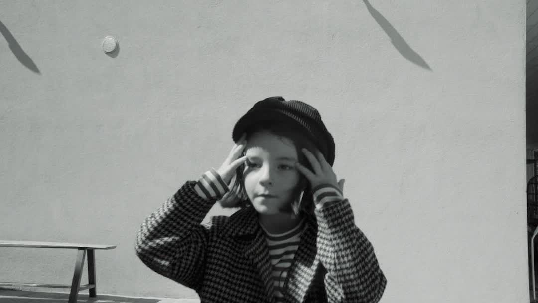 Monochrome portrait of a child in a patterned coat and hat, hands touching head, against a plain wall with shadows.