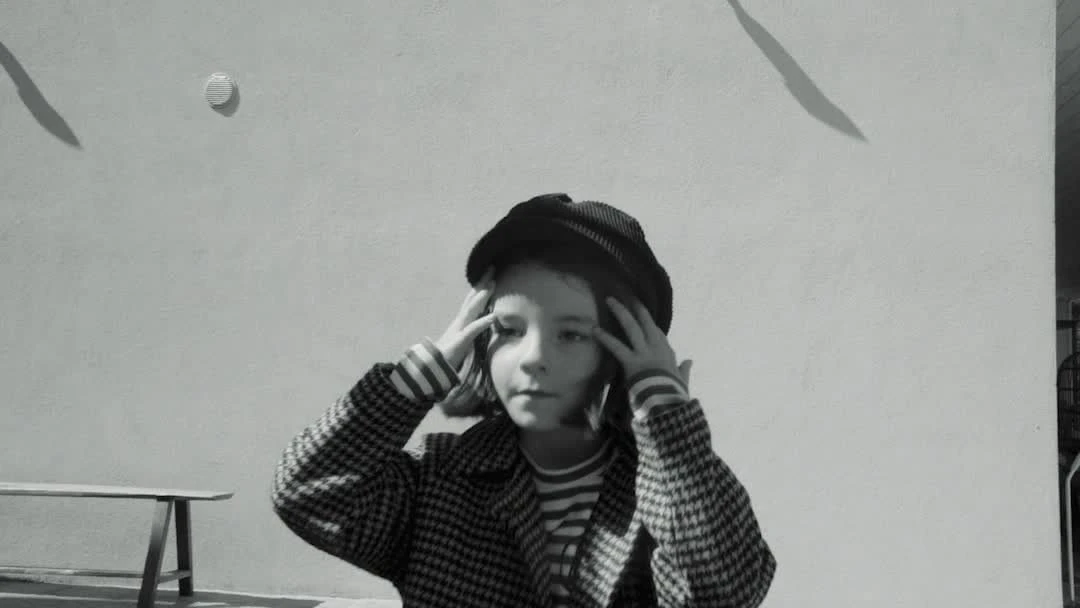Monochrome portrait of a child in a patterned coat and hat, hands touching head, against a plain wall with shadows.