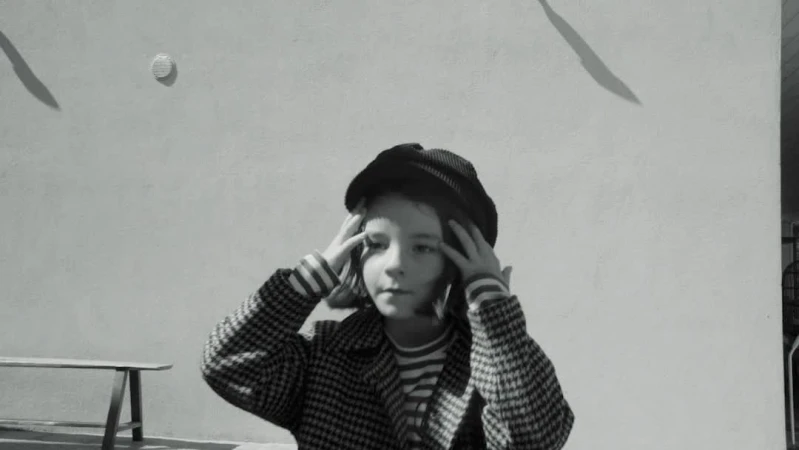 Monochrome portrait of a child in a patterned coat and hat, hands touching head, against a plain wall with shadows.