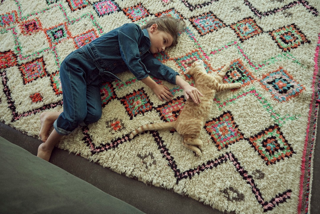A young child in a denim outfit lies on an ornate rug next to a ginger cat who is lying on its back and being stroked by the child.