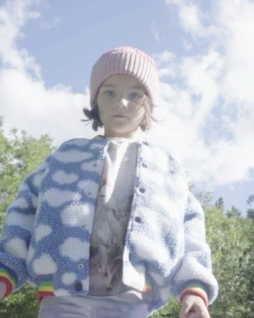 Child wearing a pink beanie and a blue cloud patterned jacket photographed outdoors against a blue sky with scattered clouds.