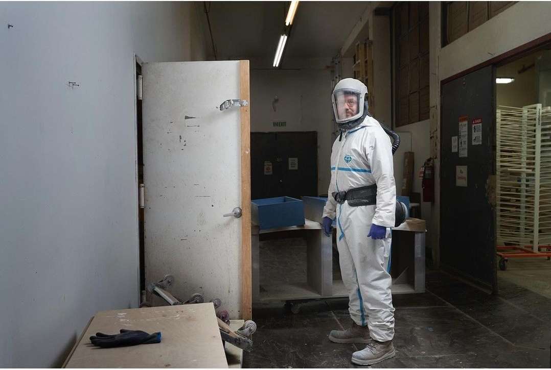 Man in hazmat suit and full face shield stands in industrial workspace with closed door and shelving in the background.