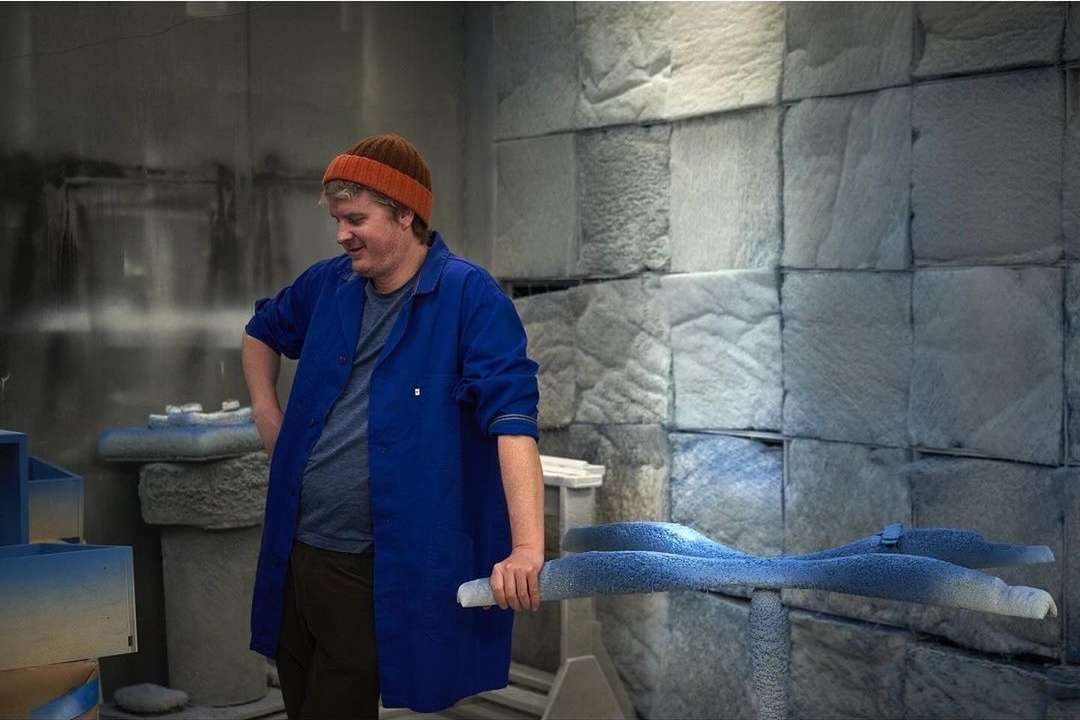 Adult man in blue coat and orange hat in a workshop holding a blue object in his hand with stone textured wall in the background.