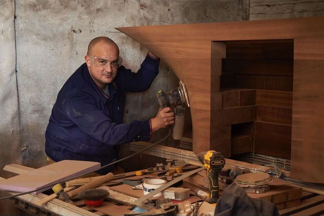 Carpenter smoothing wood with a sander, wearing safety glasses, indoors with woodworking tools on a table, against a textured wall.