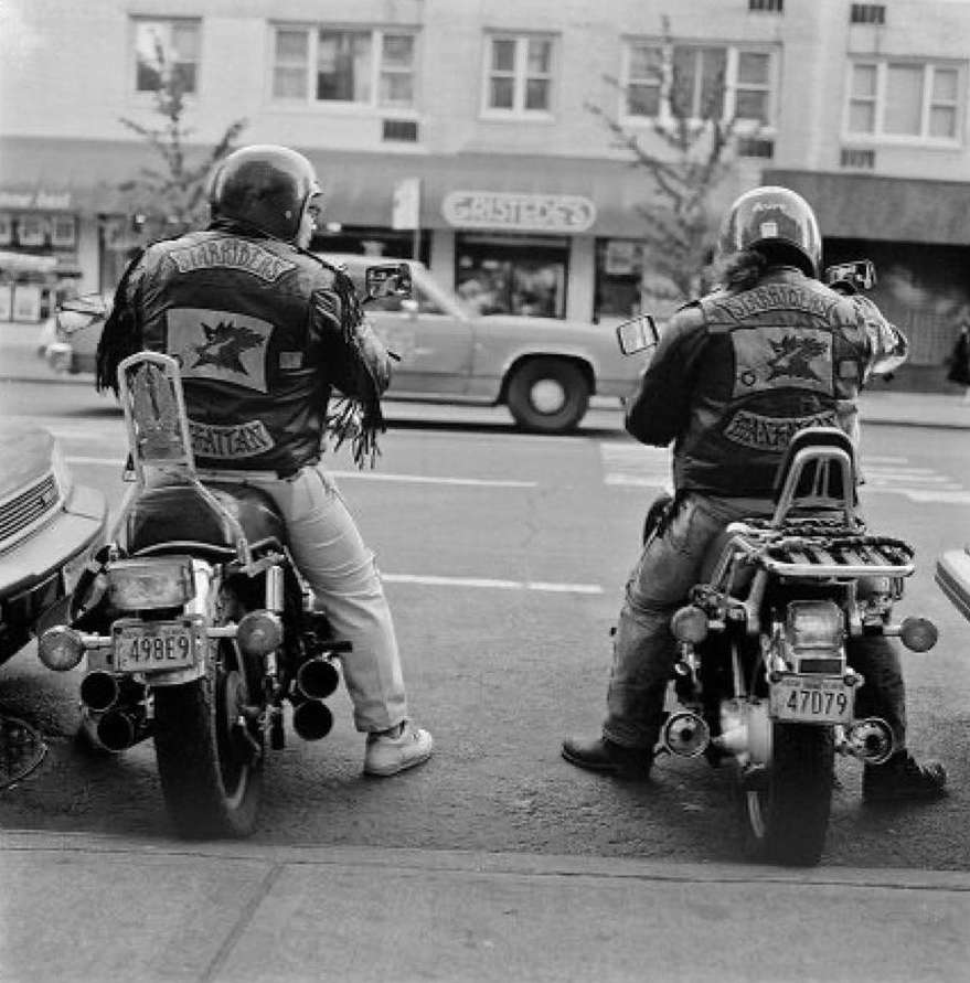 Two motorcyclists with helmets and leather jackets, parked on motorcycles in a city street with buildings and a car in the background.
