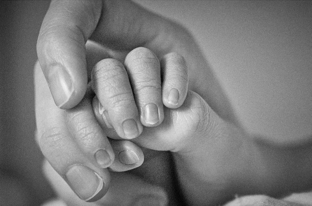 Close-up black and white photograph of an adult hand holding a baby's hand, showing the size contrast and delicate nature of the baby's fingers.