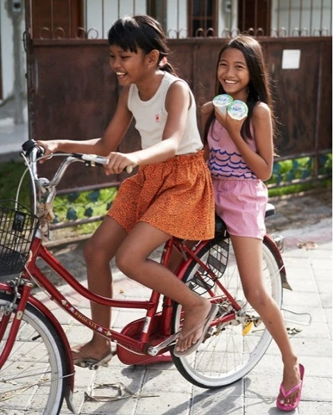 Two smiling Asian girls on a red bicycle, one riding and one standing behind holding two small containers outside a house with a gate.