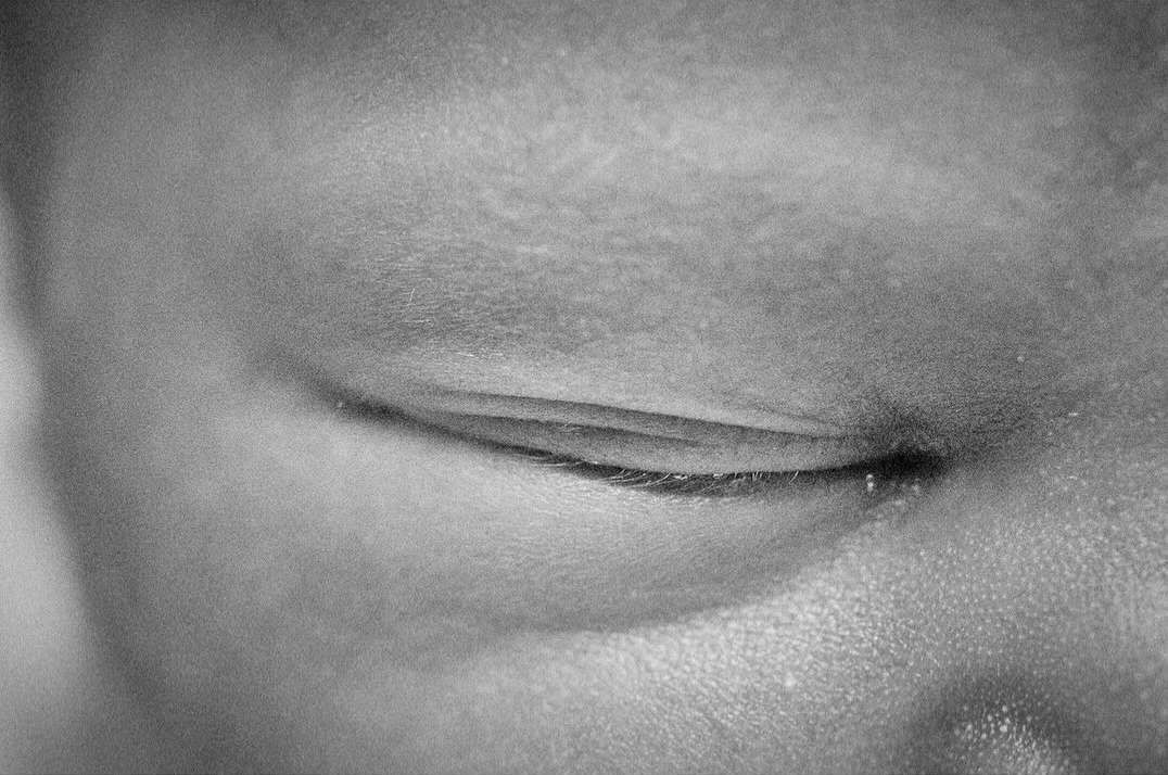 Macro shot in black and white of a closed human eye, showing eyelashes and skin texture, with soft, diffused lighting.