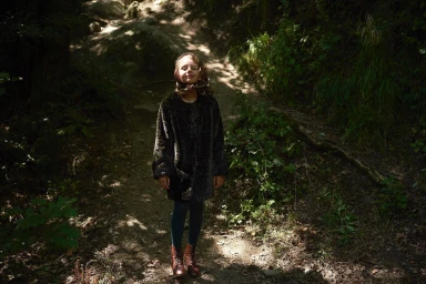 A young girl wearing a dark furry coat, headscarf, tights, and brown boots stands on a woodland path under natural light.