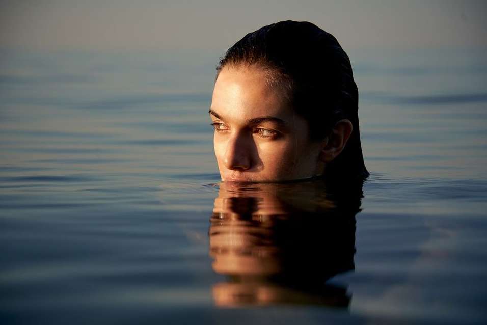 Woman partially submerged in water, facing left with light on face. Body of water extends to the horizon under a clear sky.