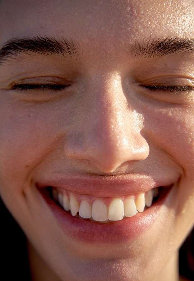 Close-up portrait of a smiling woman with closed eyes, fair skin, and white teeth, captured in bright sunlight. Beauty concept.