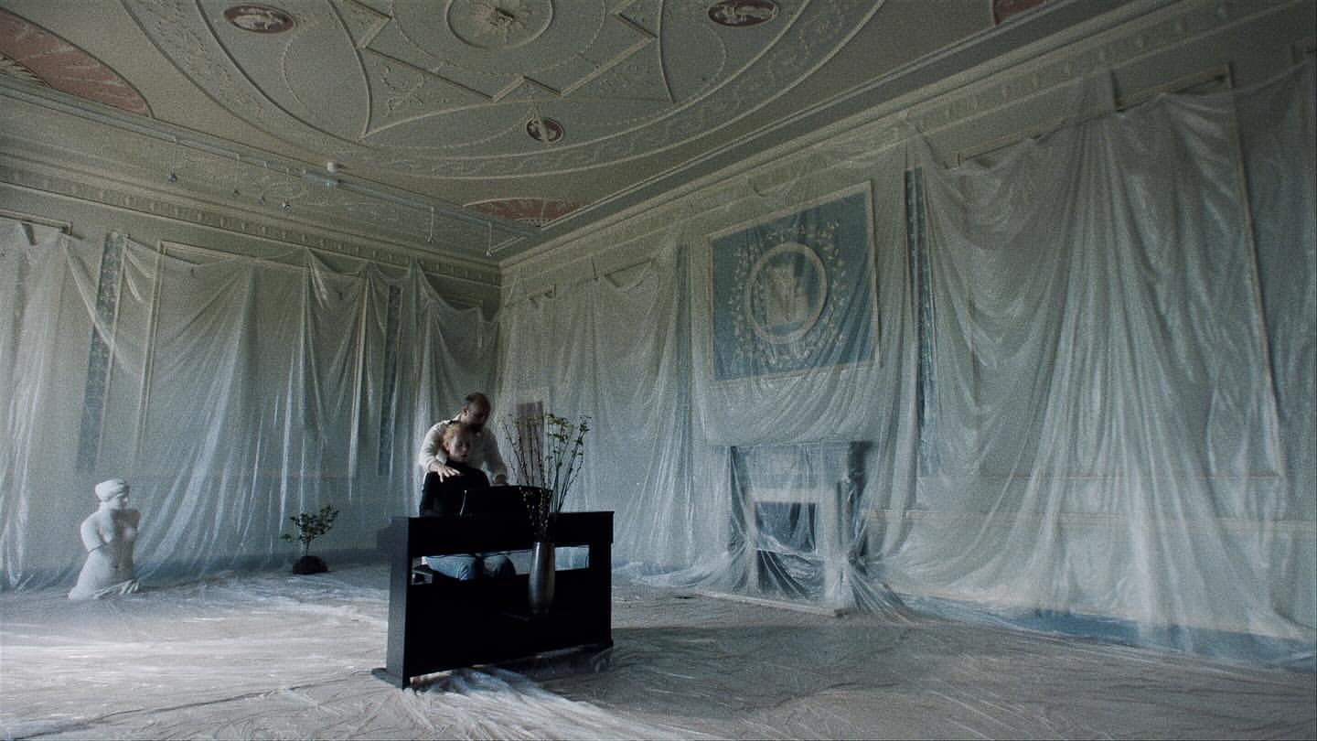 A man and woman are in an indoor setting with furniture partially covered in plastic sheeting, with classical design elements.