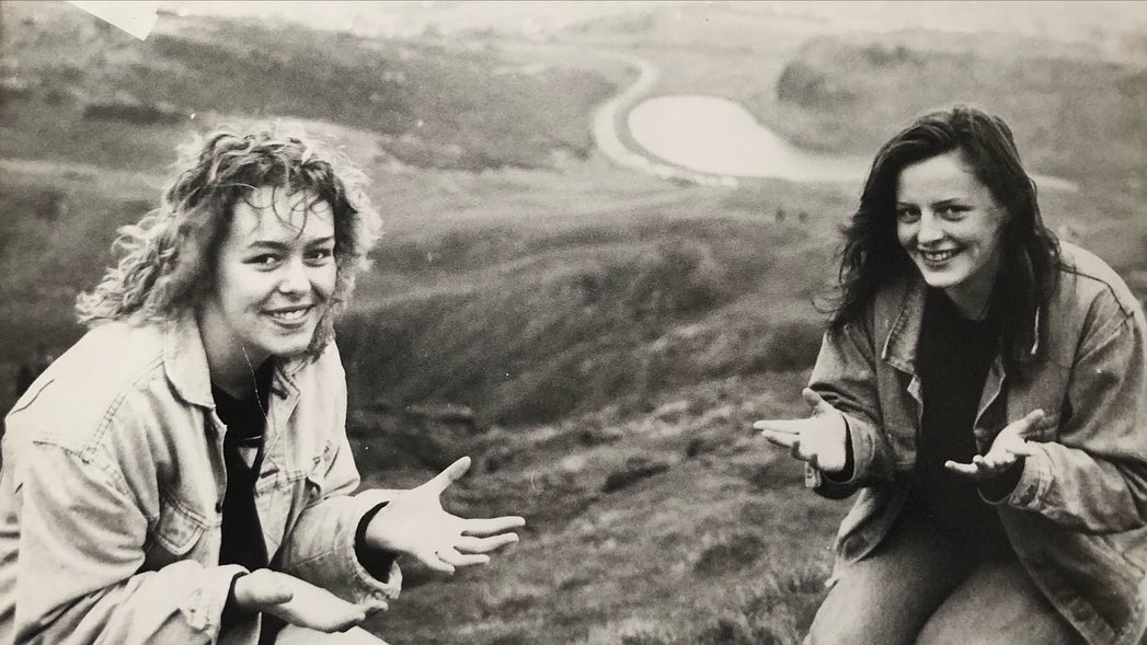 Two smiling women pose outdoors against a hillside backdrop featuring a winding path and pond, captured in a black-and-white photograph.