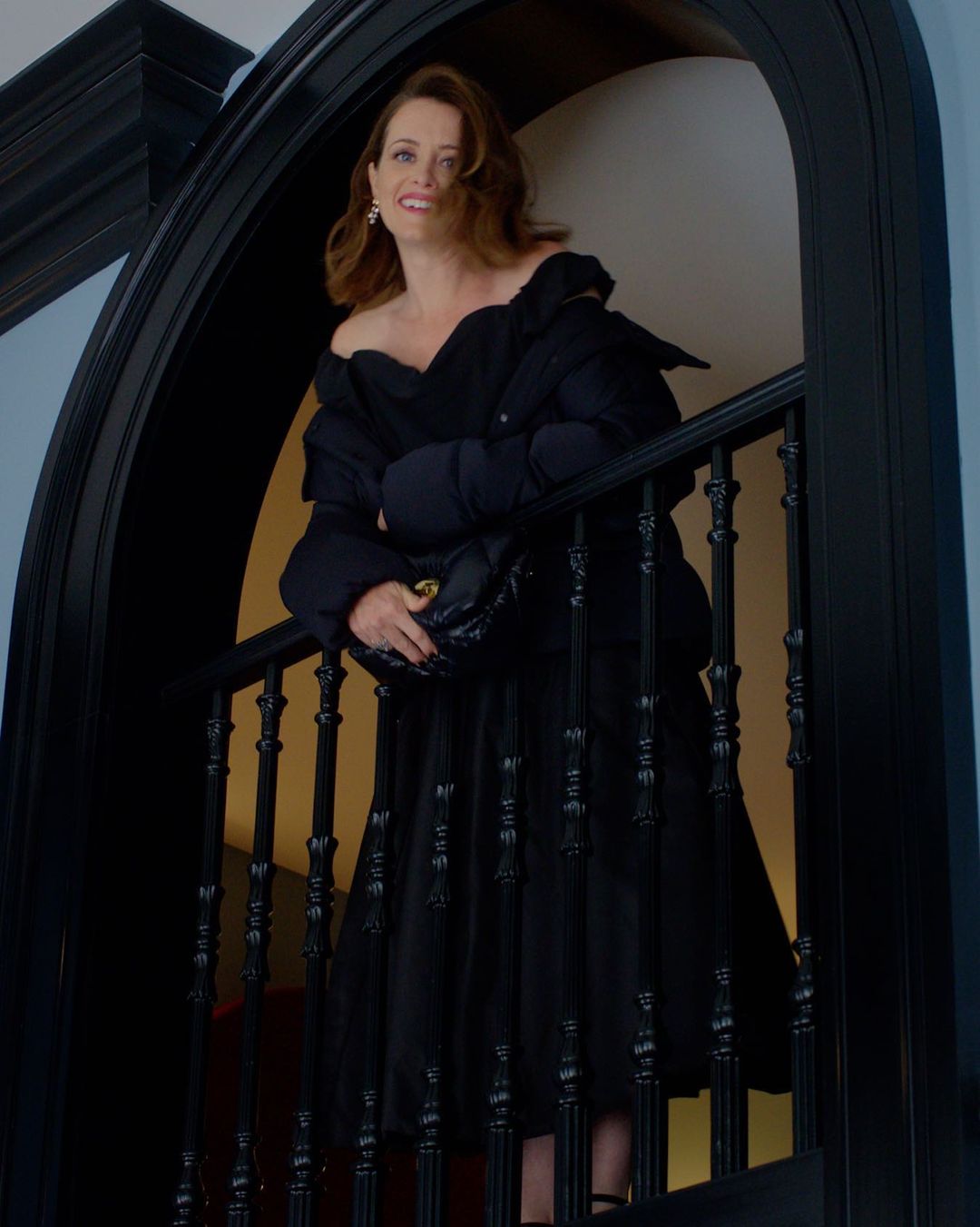 Woman in black dress and jacket standing on a staircase with black banisters, holding a black purse, smiling and looking up.
