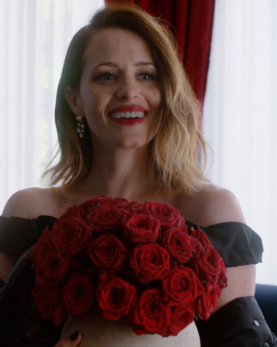 Woman with red lipstick holds a bouquet of red roses in a bright room. She is smiling while wearing a dark off-the-shoulder dress.