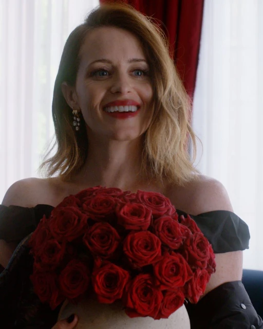 Woman with red lipstick holds a bouquet of red roses in a bright room. She is smiling while wearing a dark off-the-shoulder dress.