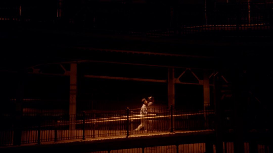 A man and a child are seen jogging at night on an elevated platform illuminated by dim yellow lighting, with a dark, urban backdrop.