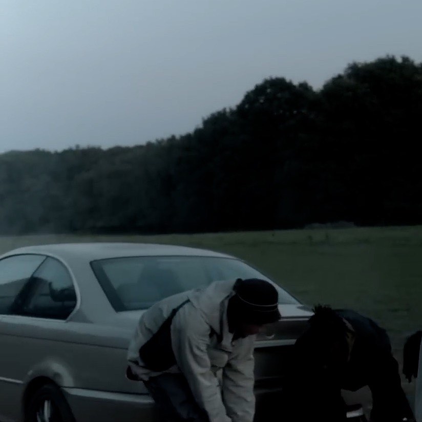 Two people near a parked silver car in a rural field with forest backdrop under a cloudy sky; one person appears to be tying a shoe.