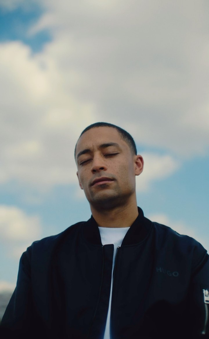 Man with closed eyes in black bomber jacket poses against a blue sky with white clouds. Outdoor portrait photography. Serene mood.