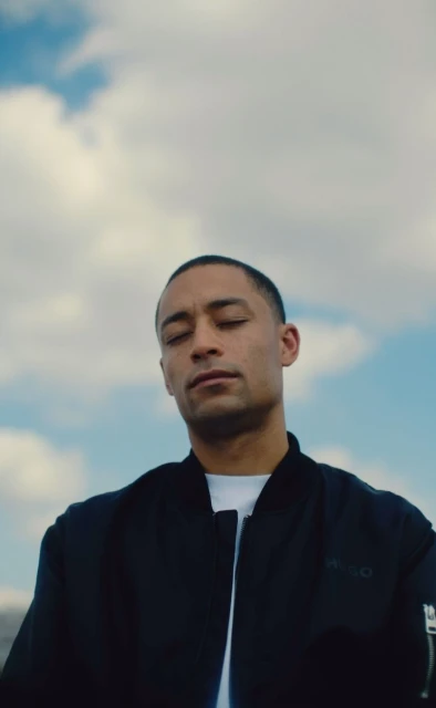 Man with closed eyes in black bomber jacket poses against a blue sky with white clouds. Outdoor portrait photography. Serene mood.