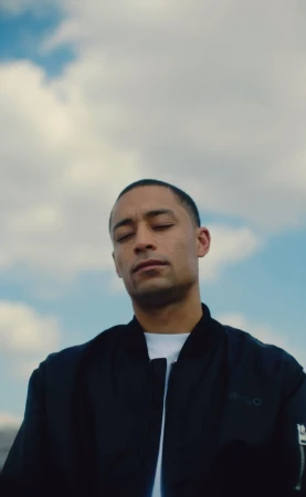 Man with closed eyes in black bomber jacket poses against a blue sky with white clouds. Outdoor portrait photography. Serene mood.