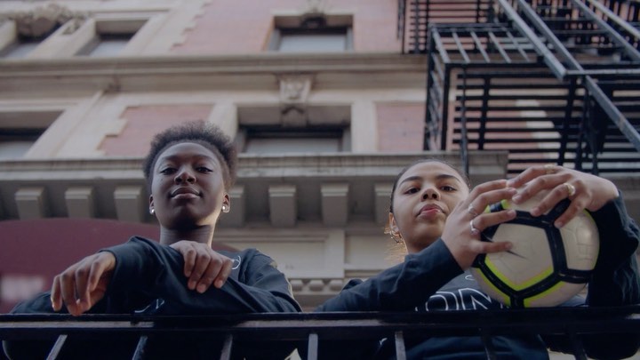 Two young women pose on a balcony overlooking a street, one holding a soccer ball. An urban building and fire escape form the backdrop.