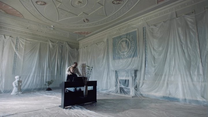 Couple in a light-filled room covered with plastic, standing behind a black piano with a vase of flowers. Renovations, fine art.
