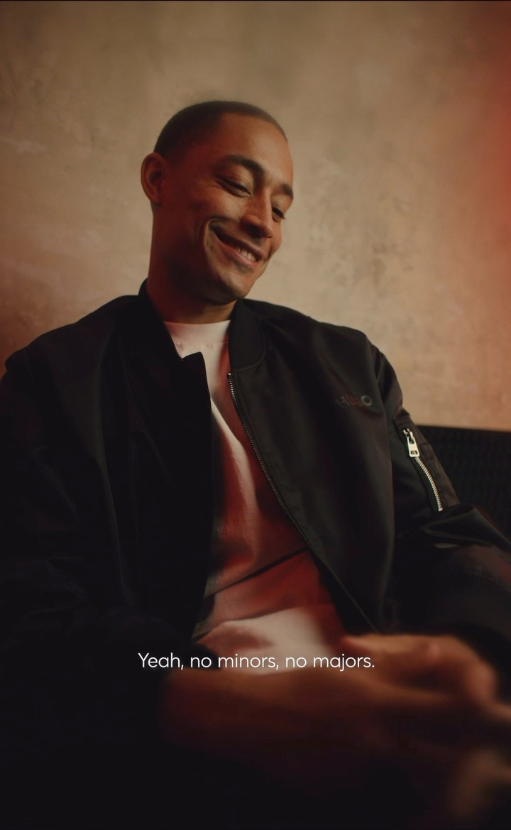 A smiling man is indoors, wearing a black jacket and white shirt, illuminated by warm lighting against a textured background, captured at eye-level.