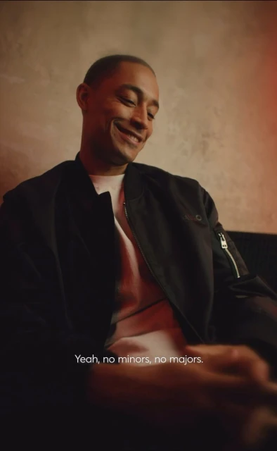 A smiling man is indoors, wearing a black jacket and white shirt, illuminated by warm lighting against a textured background, captured at eye-level.