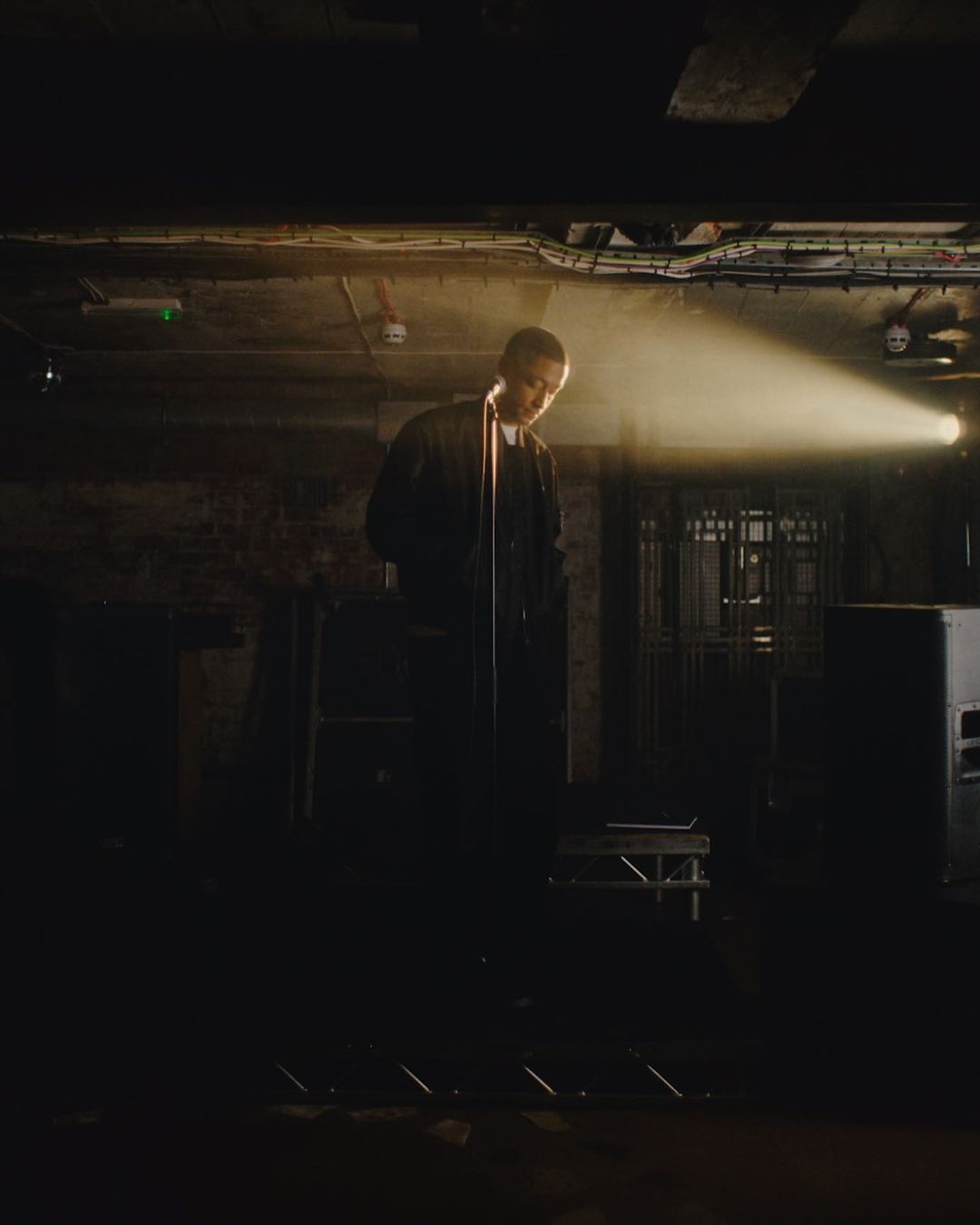Male rapper performing live on stage in dim underground music venue with brick wall and yellow spotlight; dark clothing and equipment.