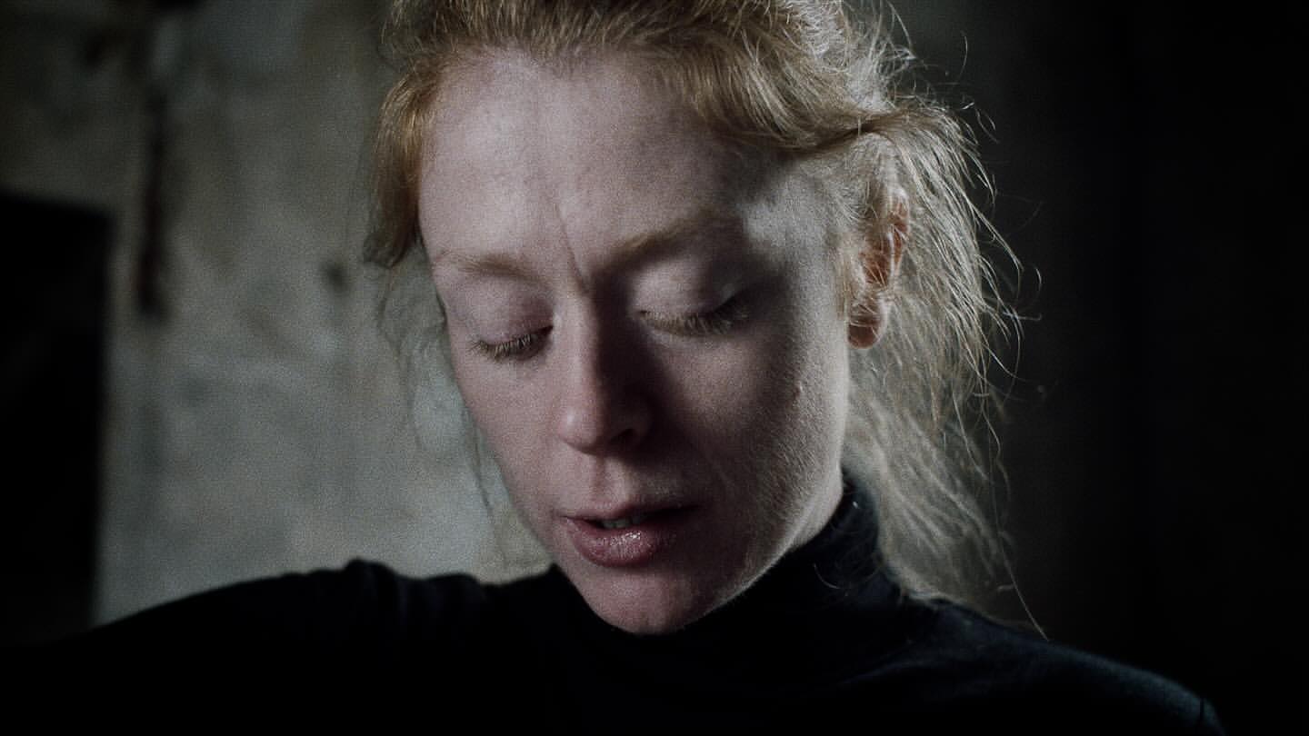 Close-up portrait of a fair-skinned adult female with closed eyes and light red hair, wearing a black turtleneck in dark lighting.