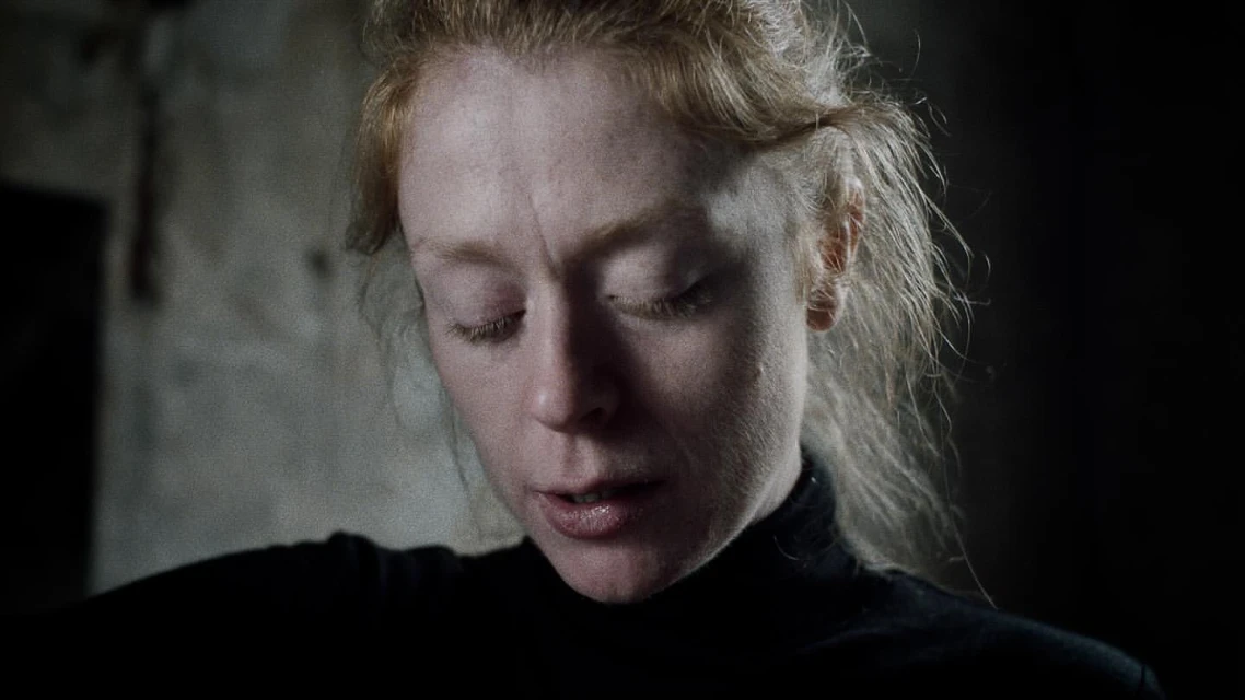 Close-up portrait of a fair-skinned adult female with closed eyes and light red hair, wearing a black turtleneck in dark lighting.