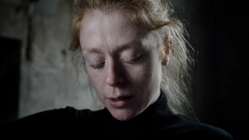 Close-up portrait of a fair-skinned adult female with closed eyes and light red hair, wearing a black turtleneck in dark lighting.