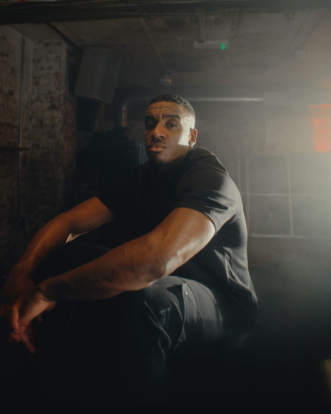 Adult male sits indoors under moody lighting wearing a black t-shirt and pants, arms resting on legs, in front of a textured wall.