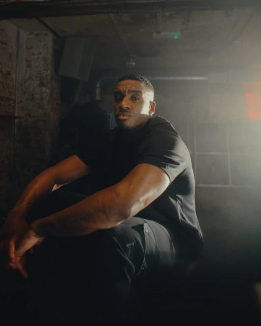 Adult male sits indoors under moody lighting wearing a black t-shirt and pants, arms resting on legs, in front of a textured wall.