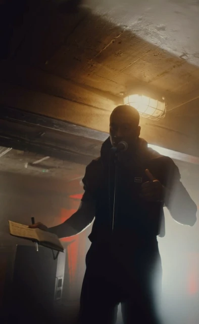 Silhouette of adult man wearing a jacket standing in front of a microphone holding a notebook in an indoor environment with lights.
