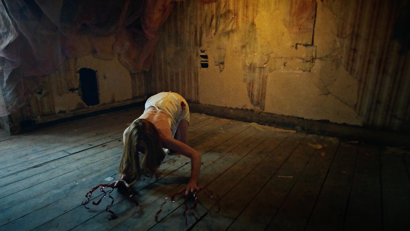 A woman with blood-covered hands and bare feet crawls on all fours in an aged room, creating a chilling and dramatic scene.