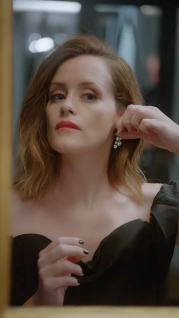 Woman in black dress adjusts pearl earrings indoors with soft lighting and mirror reflection, capturing a moment of personal grooming.