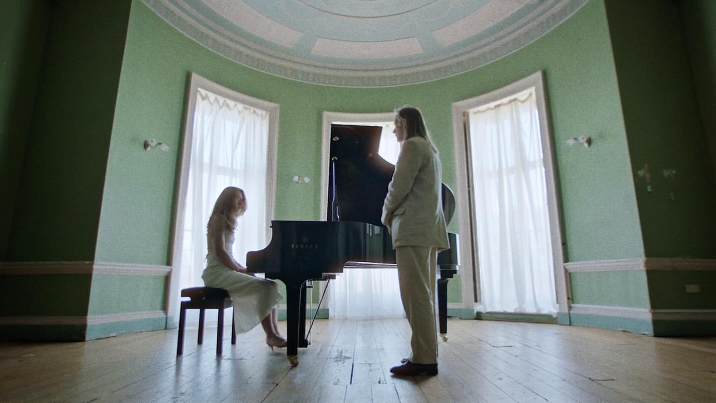 A woman plays a black grand piano while a man watches in a large room with light green walls and tall windows with white curtains.