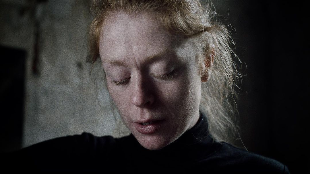 Close-up portrait of a redhead woman in a black turtleneck looking down, with a shallow depth of field and dark, muted tones.
