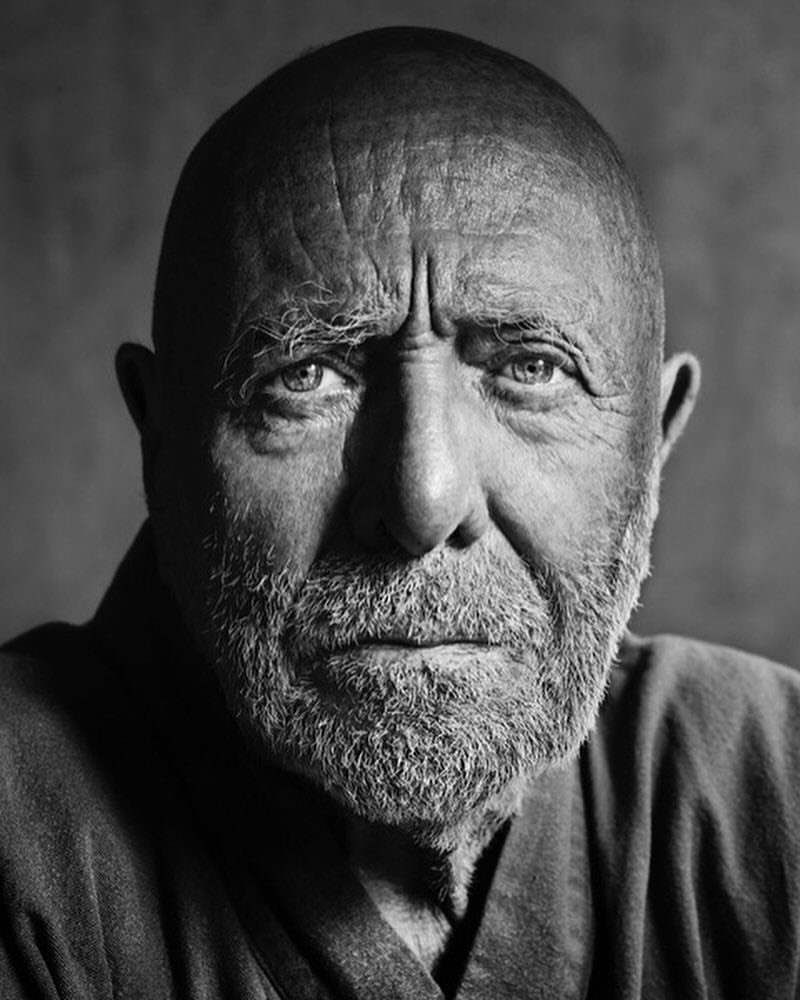 Black and white portrait of a mature adult man with a shaved head and full beard, looking directly at the camera with a serious expression.